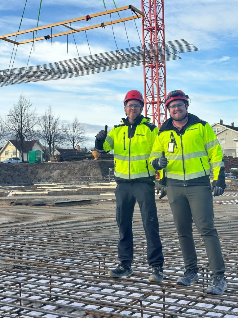 To menn på byggeplass, verneklær, hjelm, tommel opp, heiskran og armeringsjern, bunnplate i bakgrunnen, sol, lettskya