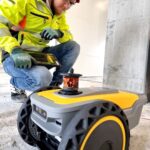 Byggeplass, mann i verneutstyr sitter på huk, ser på en robot på et tomt betonggulv